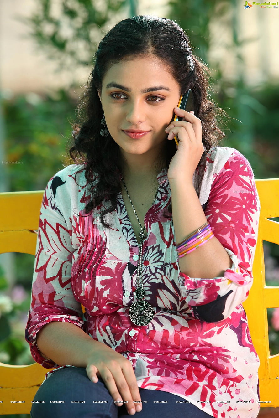 Nithya Menon in a floral blouse sitting on a yellow bench talking on a mobile phone HD phone wallpaper
