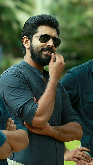 Nivin Pauly in a dark shirt and aviator sunglasses with a full beard, smiling outdoors HD phone wallpaper