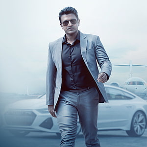 Puneeth Rajkumar in a grey suit and sunglasses walking past a luxury car at an airport HD phone wallpaper