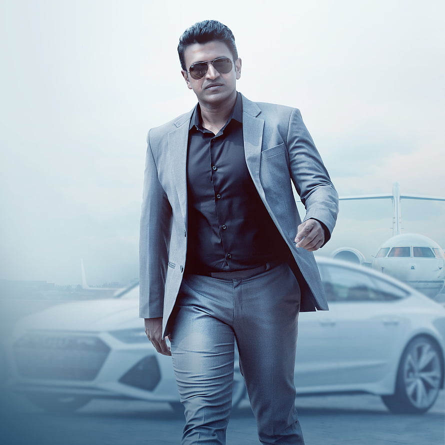 Puneeth Rajkumar in a grey suit and sunglasses walking past a luxury car at an airport HD phone wallpaper