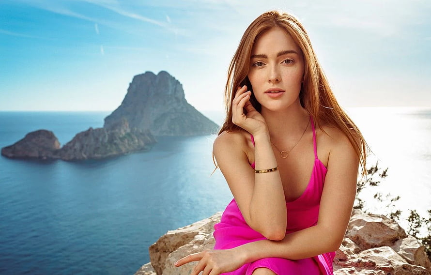 Redhead model in pink dress posing on coastal cliff with ocean and rocky island backdrop HD wallpaper