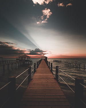 Dramatic sunset over a wooden pier with sunbeams breaking through dark clouds HD phone wallpaper