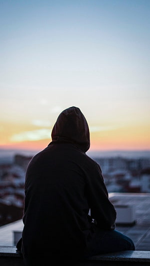 Silhouette of a hooded figure sitting on a rooftop ledge watching a colorful sunset over a city skyline HD phone wallpaper