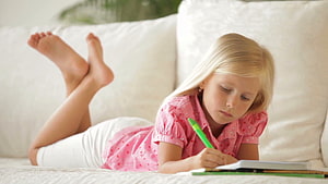 Blonde Girl Lying on White Sofa Writing in Notebook with Green Pen HD wallpaper