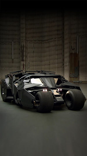 The Dark Knight Batmobile in a dim industrial hangar with glowing headlights and rugged tires HD phone wallpaper