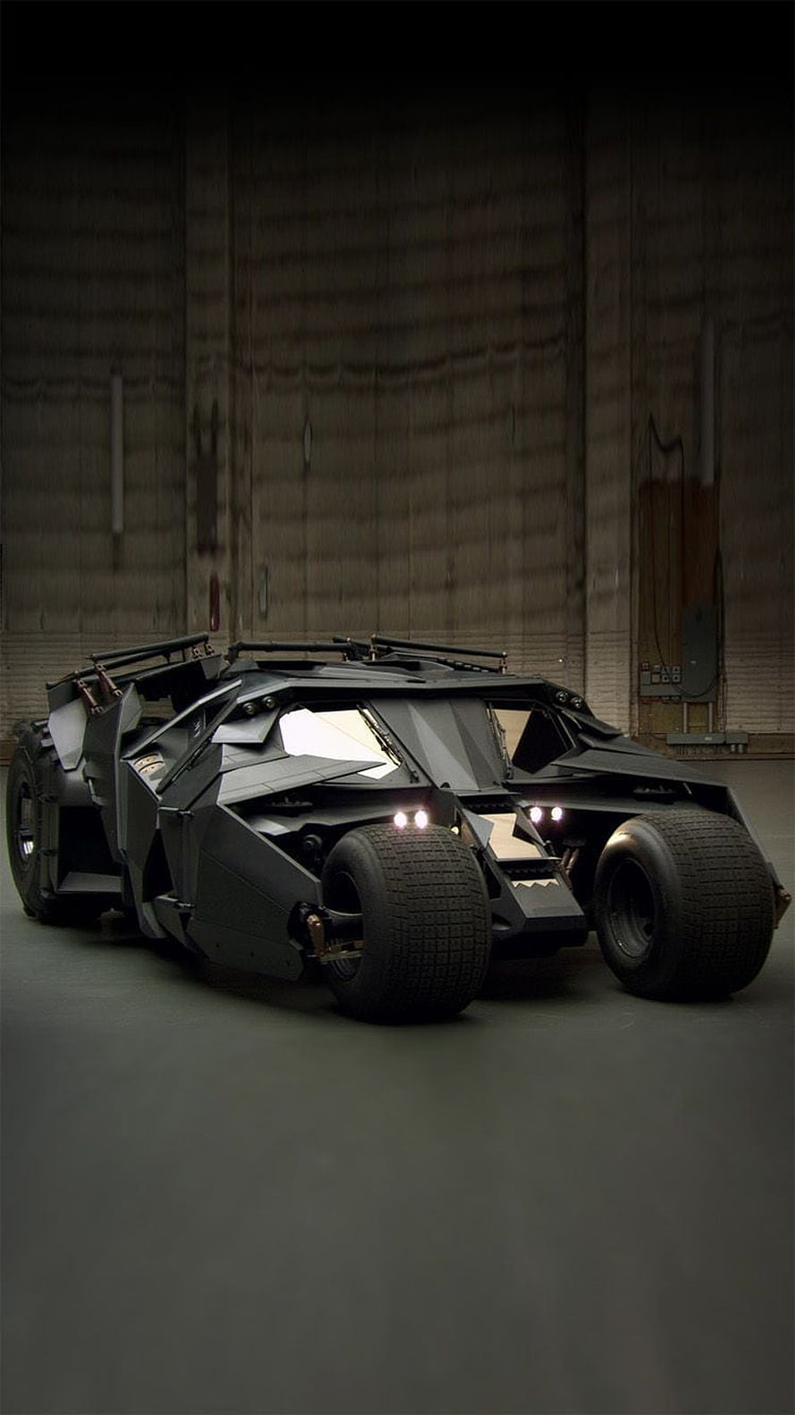 The Dark Knight Batmobile in a dim industrial hangar with glowing headlights and rugged tires HD phone wallpaper