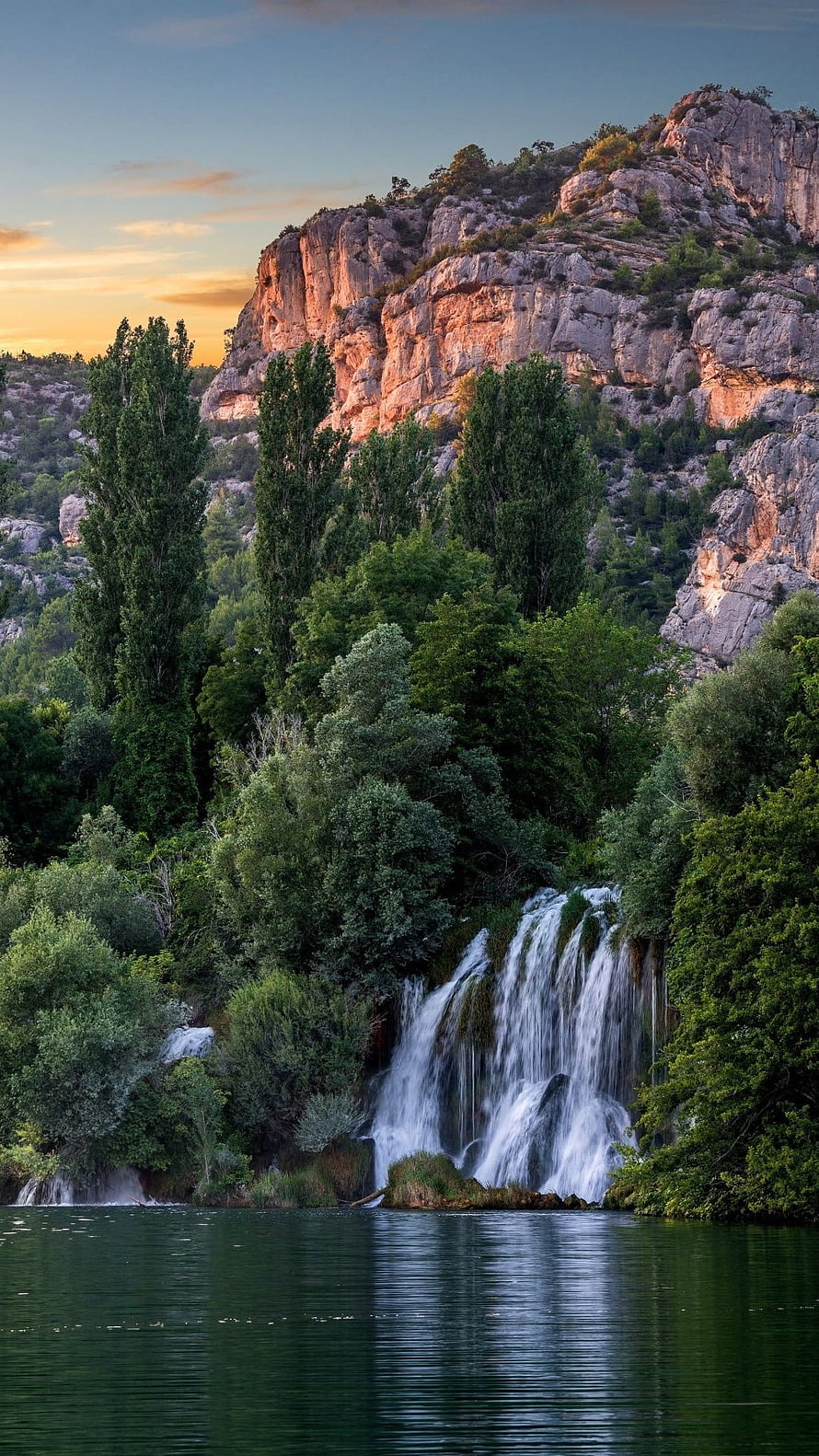 Serene Waterfall Cascading Into a Calm Lake Beneath a Rocky Cliff at Golden Hour HD phone wallpaper
