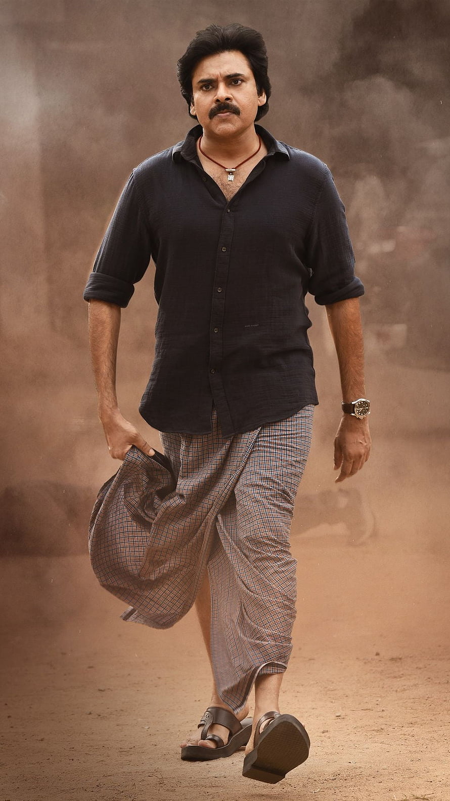 Pawan Kalyan in a black shirt and checkered lungi walking through a dusty rural landscape HD phone wallpaper