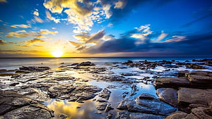 Golden sunset over a rocky ocean shoreline with dramatic clouds and water reflections HD wallpaper