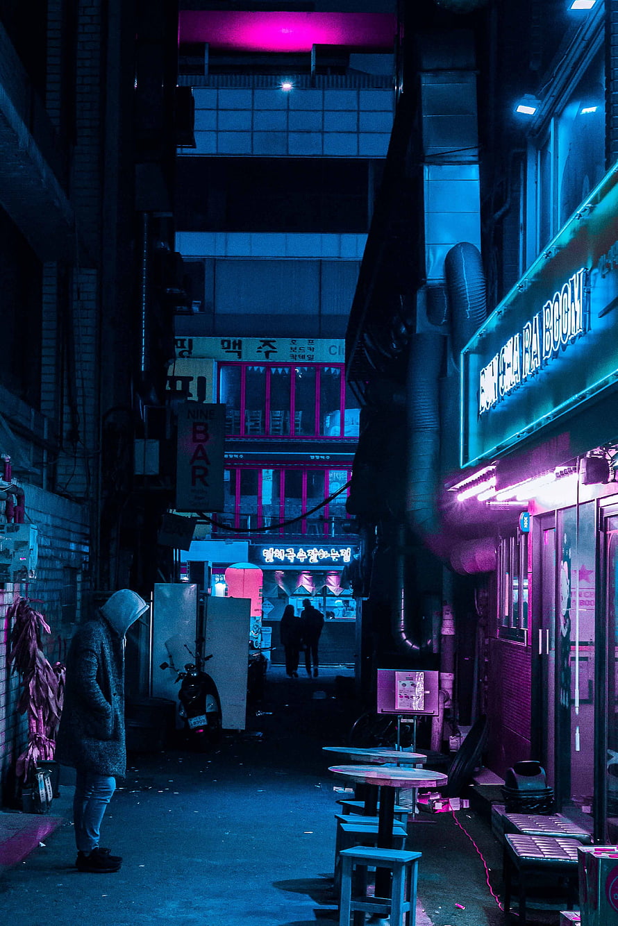 Neon-lit urban alleyway at night with glowing signs and a lone figure in a hooded coat HD phone wallpaper