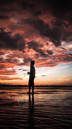 Silhouette of a lone figure standing in shallow water beneath a dramatic fiery sunset sky HD phone wallpaper