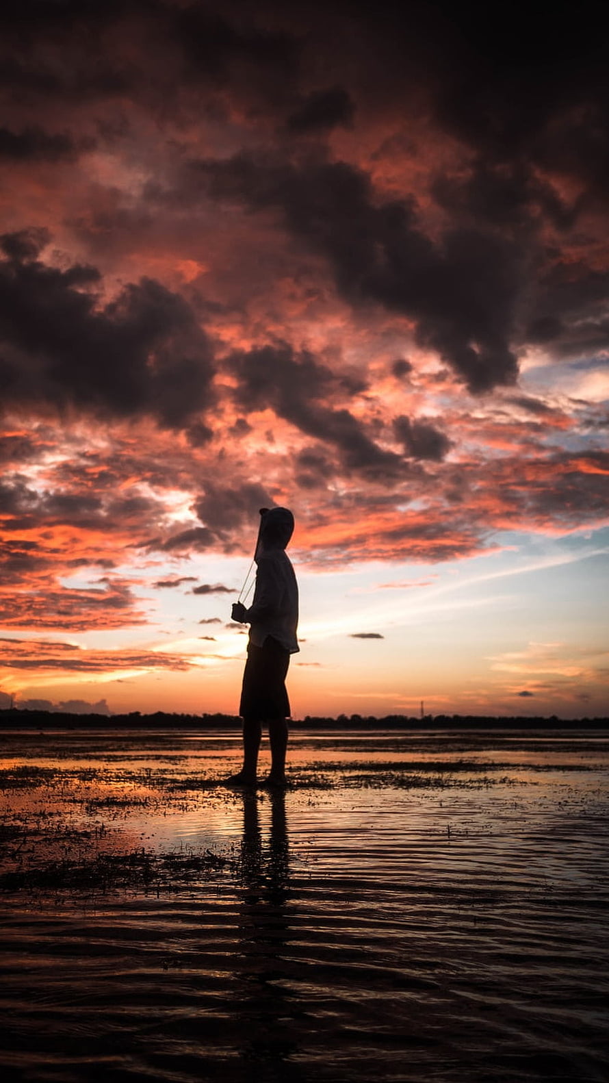 Silhouette of a lone figure standing in shallow water beneath a dramatic fiery sunset sky HD phone wallpaper