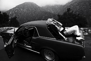 Black and White Woman Smoking on Classic Car Hood in Mountainous Landscape HD wallpaper