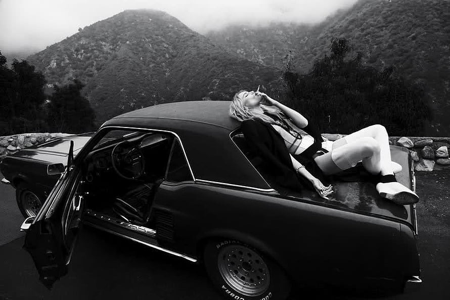Black and White Woman Smoking on Classic Car Hood in Mountainous Landscape HD wallpaper