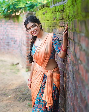 Dharsha Gupta in a vibrant blue floral lehenga and orange half saree against a rustic brick wall HD phone wallpaper