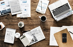 Top-down view of a cluttered office desk with laptop, calculator, business newspaper, and financial documents HD wallpaper