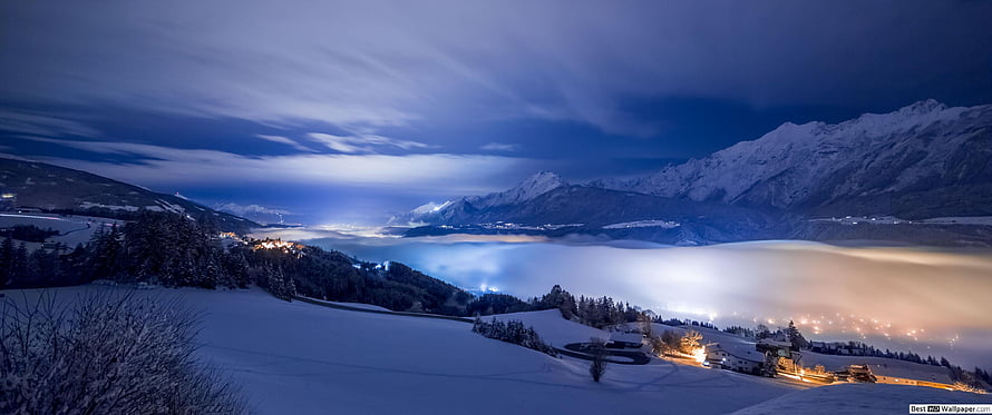 Snow-Covered Alpine Valley at Night with Glowing Village Lights and Dramatic Clouds HD wallpaper
