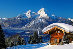 Alpine wooden chalet with snow-covered roof set against towering snowy mountain peaks and evergreen forest HD wallpaper