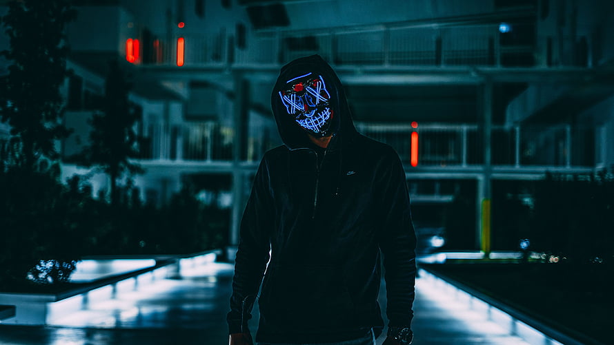 Anonymous figure in a glowing LED mask and dark hoodie standing in a neon-lit urban corridor at night HD wallpaper
