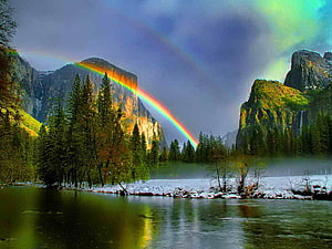 Vibrant rainbow arching over a serene mountain lake with evergreen forest and snow-dusted banks HD wallpaper