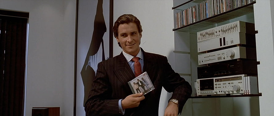 Patrick Bateman in a pinstripe suit holding a cassette tape in a modern room with audio equipment HD wallpaper