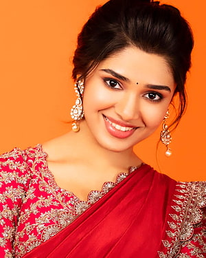 Kriti Shetty in a red embroidered sari with gold jewelry against an orange backdrop HD phone wallpaper