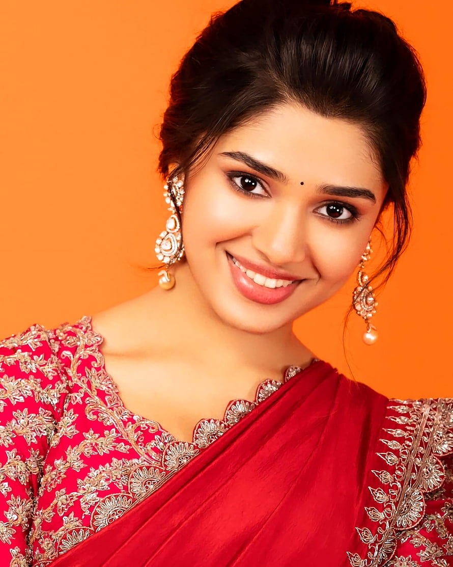 Kriti Shetty in a red embroidered sari with gold jewelry against an orange backdrop HD phone wallpaper