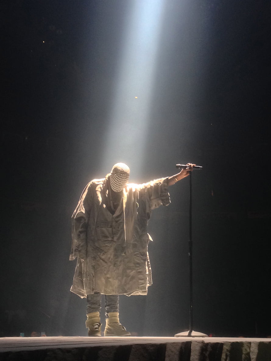 Kanye West performing on stage in a chainmail mask under a dramatic spotlight HD phone wallpaper