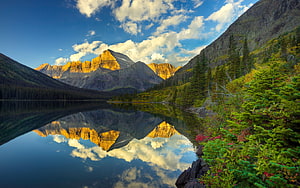 Golden mountain peaks reflected in a calm alpine lake surrounded by lush green forest and blue sky HD wallpaper