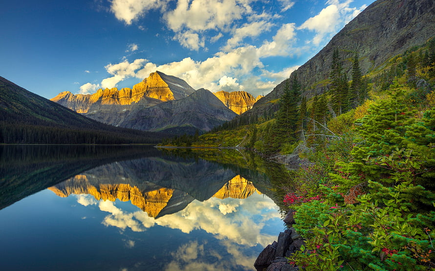 Golden mountain peaks reflected in a calm alpine lake surrounded by lush green forest and blue sky HD wallpaper