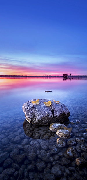 Serene Lake Sunset with Autumn Leaves on Rocks and Distant Pier HD phone wallpaper