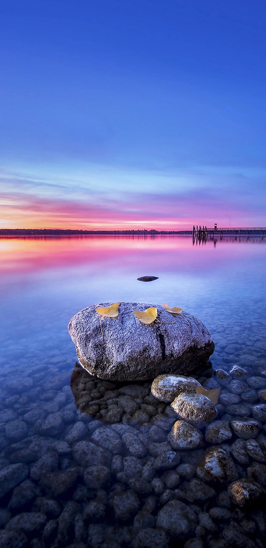 Serene Lake Sunset with Autumn Leaves on Rocks and Distant Pier HD phone wallpaper