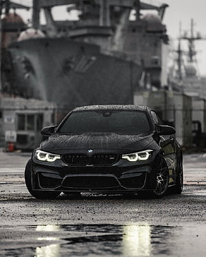 Black BMW M4 Coupe Parked on Wet Dockyard Ground with Naval Vessel Background HD phone wallpaper