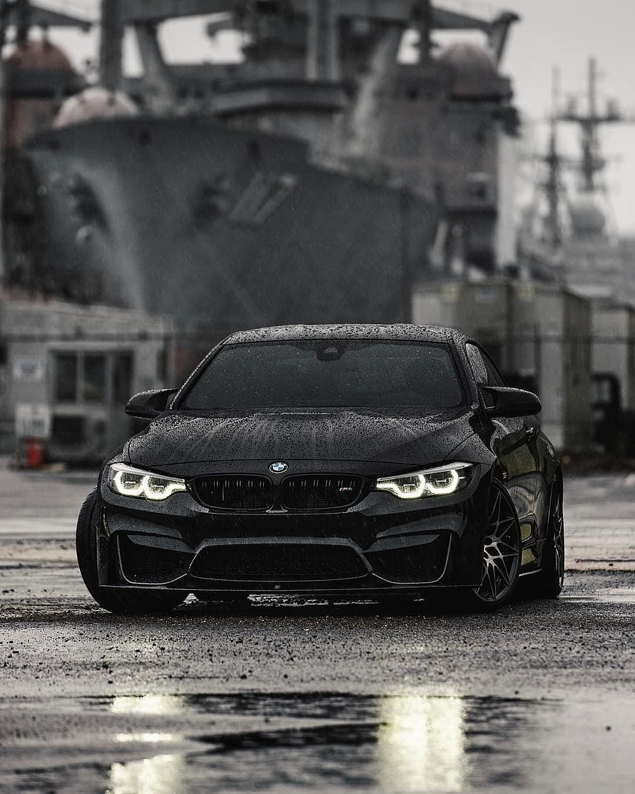 Black BMW M4 Coupe Parked on Wet Dockyard Ground with Naval Vessel Background HD phone wallpaper