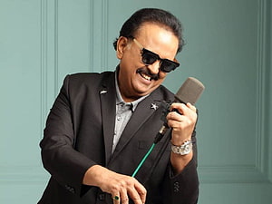 S. P. Balasubrahmanyam in a dark suit and sunglasses holding a microphone against a teal wall HD wallpaper