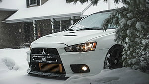 White Mitsubishi Lancer Evolution X parked in deep snow with falling flakes and pine branches HD wallpaper