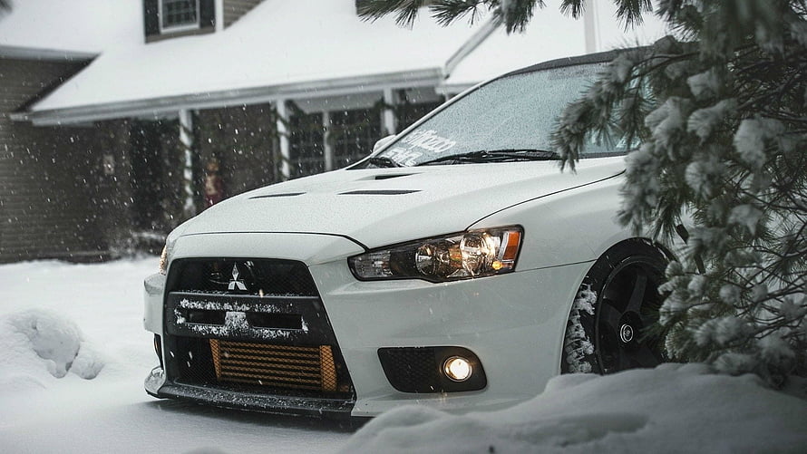 White Mitsubishi Lancer Evolution X parked in deep snow with falling flakes and pine branches HD wallpaper