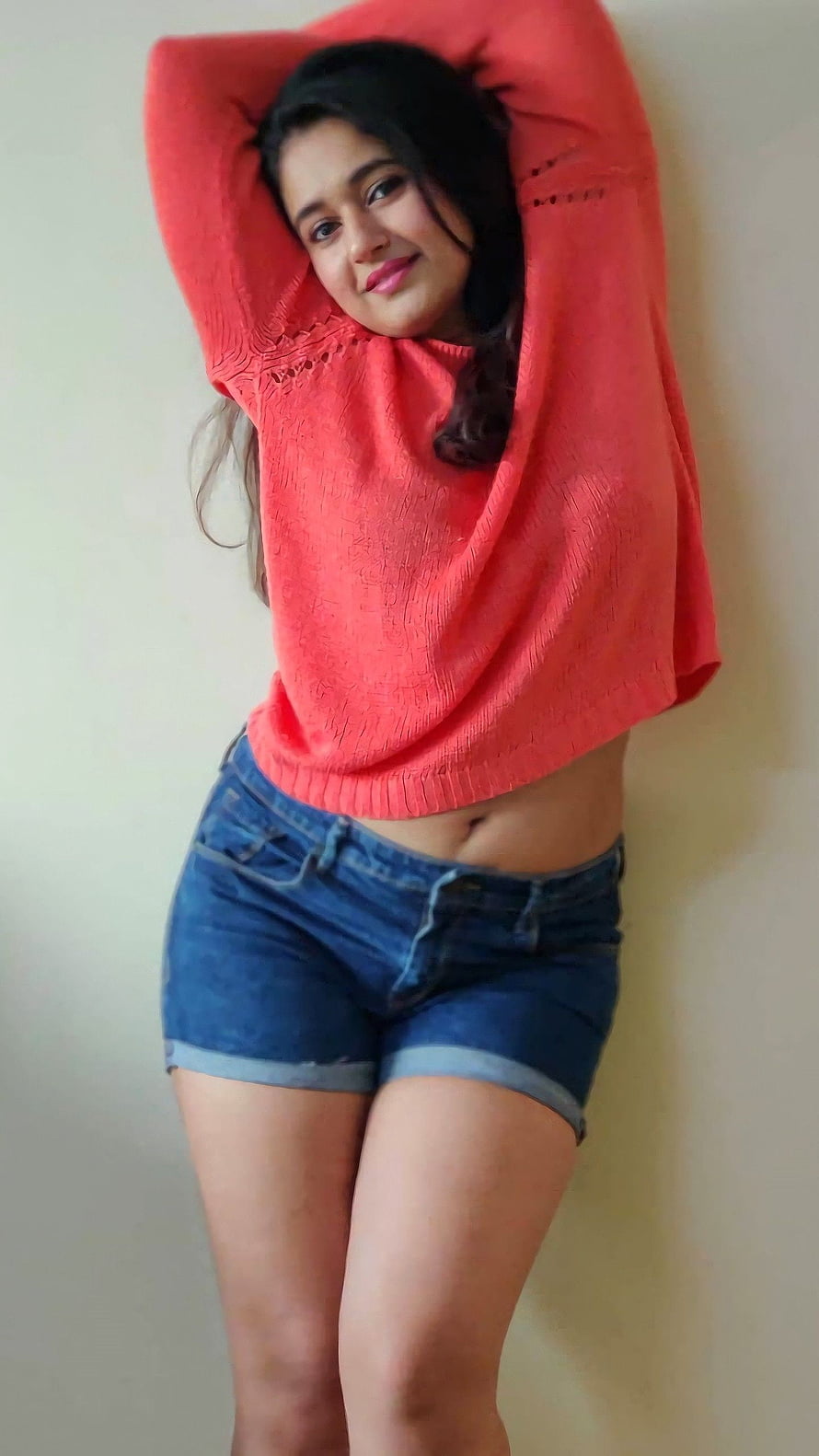 Poonam Bajwa in a coral knit top and denim shorts posing against a plain wall HD phone wallpaper