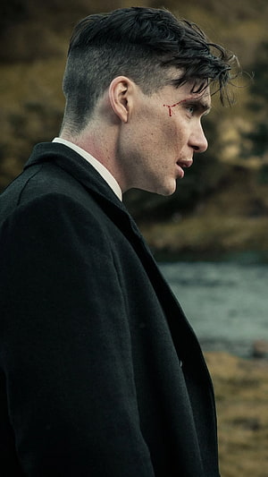 Tommy Shelby in a black overcoat with a cut on his face, set against a moody outdoor backdrop HD phone wallpaper