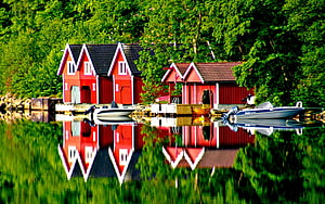 Vibrant Red Boathouses Mirrored in a Calm Green Lake with Docked Motorboats HD wallpaper