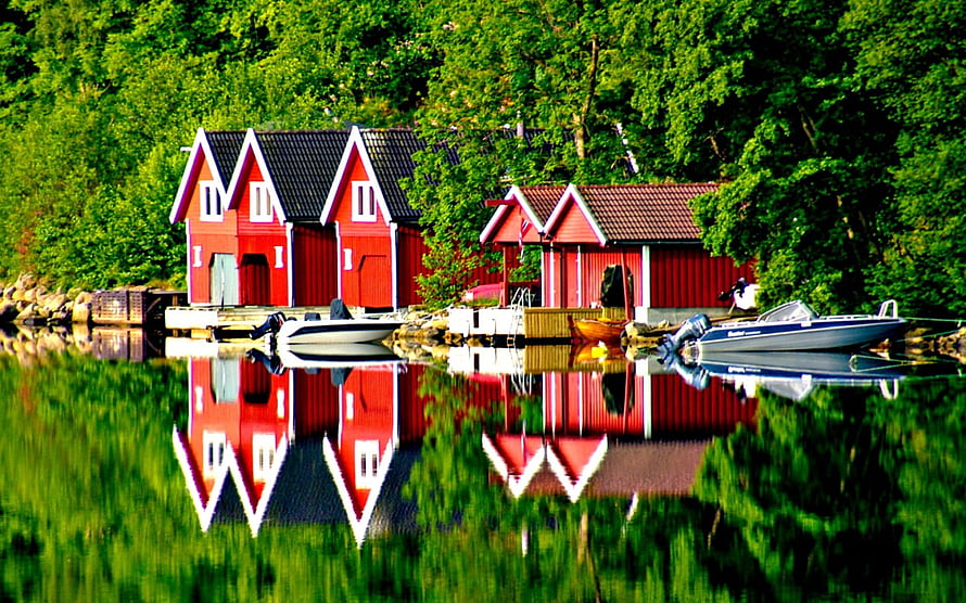 Vibrant Red Boathouses Mirrored in a Calm Green Lake with Docked Motorboats HD wallpaper