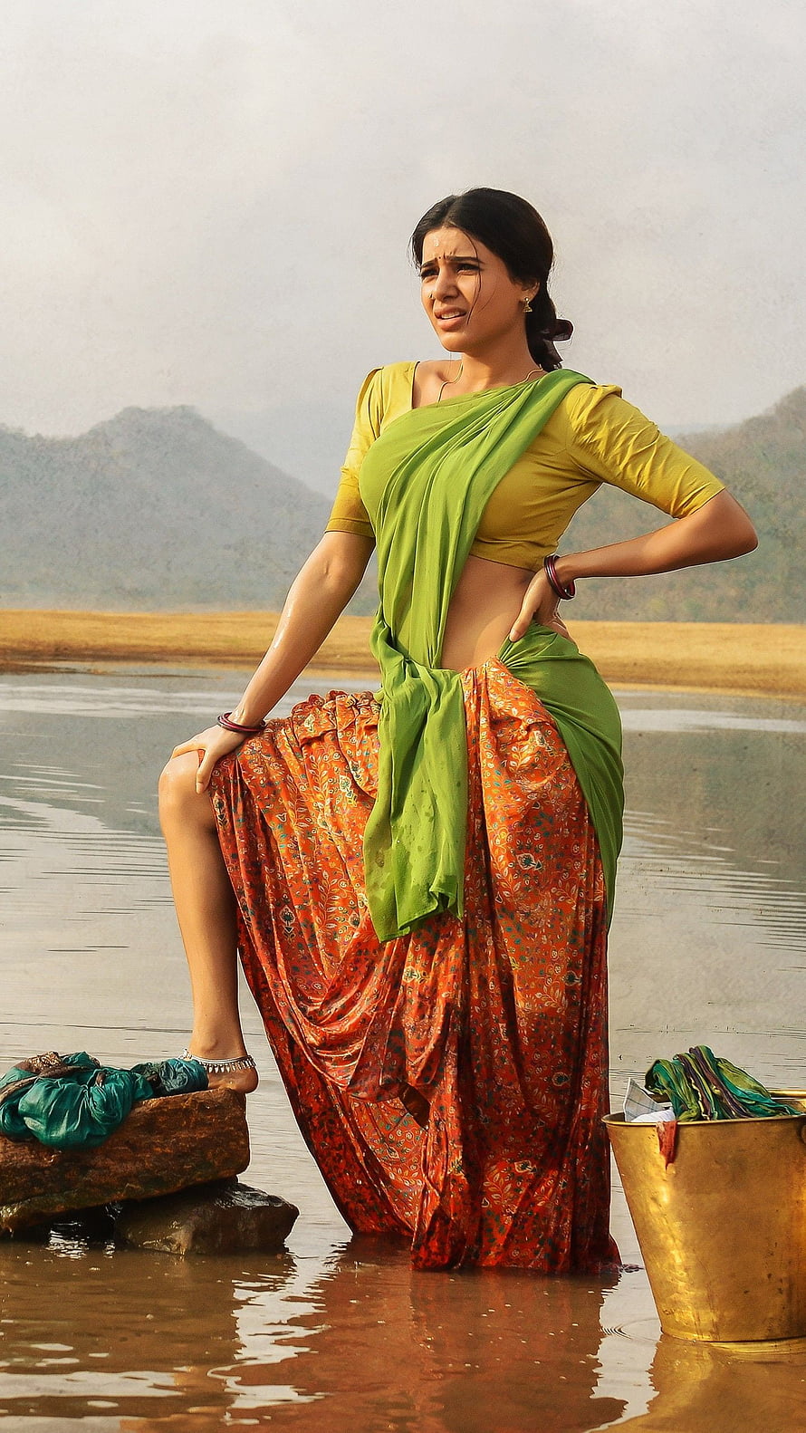 Samantha in a green and orange saree standing in a river with a brass bucket and mountains in the background HD phone wallpaper