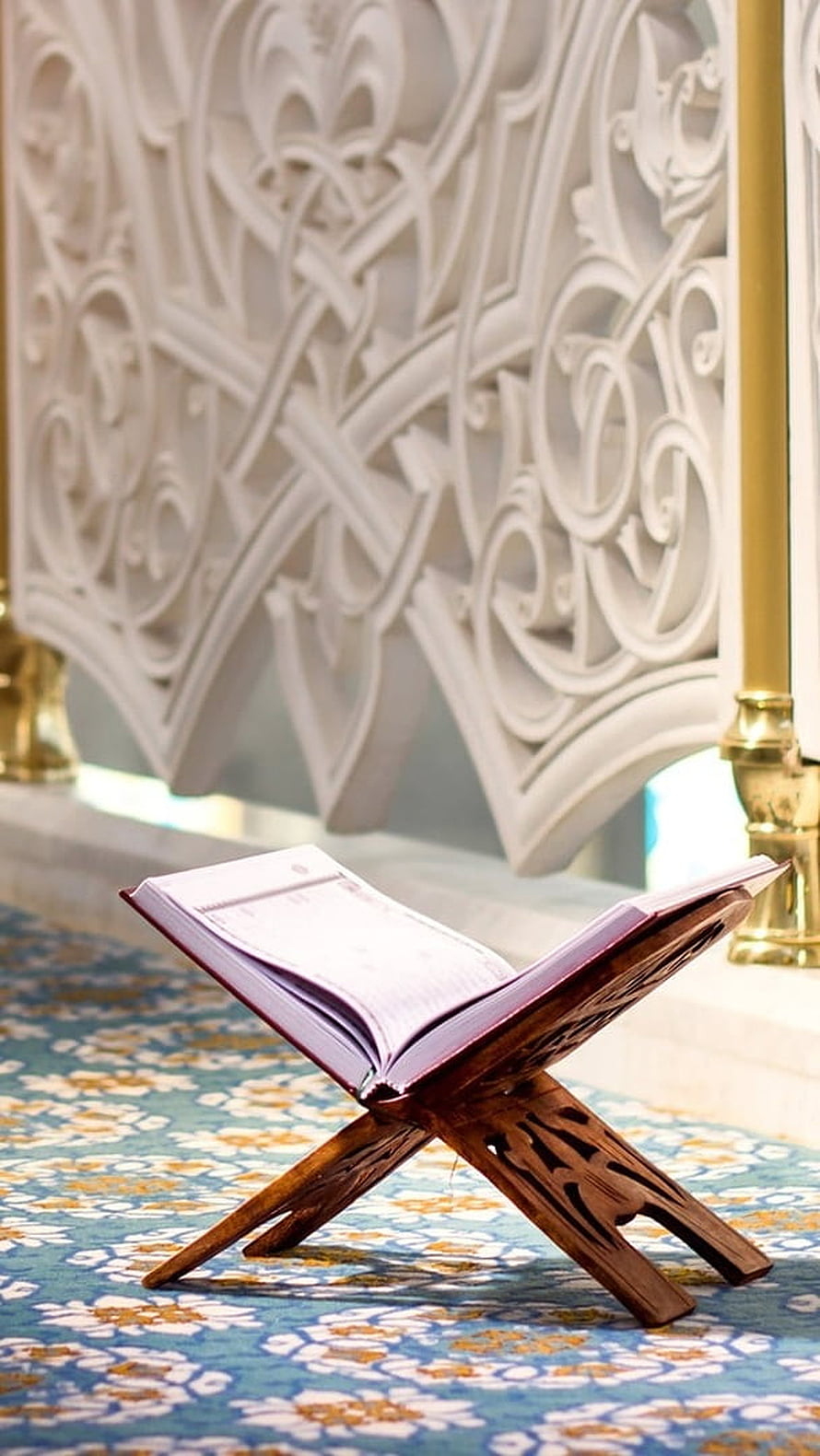 Open Quran on a wooden stand inside a mosque with ornate white lattice screen and patterned carpet HD phone wallpaper