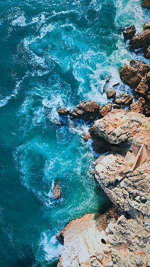 Aerial View of Turquoise Ocean Waves Crashing Against Rugged Coastal Cliffs HD phone wallpaper