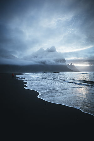 Solitary figure walking on a black sand beach beneath misty mountains and a moody overcast sky HD phone wallpaper