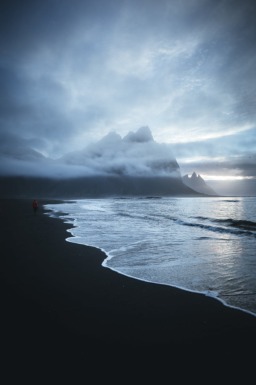 Solitary figure walking on a black sand beach beneath misty mountains and a moody overcast sky HD phone wallpaper
