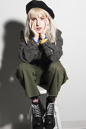 Hayley Williams in a black beret, tweed jacket, and olive trousers seated on a white stool HD phone wallpaper