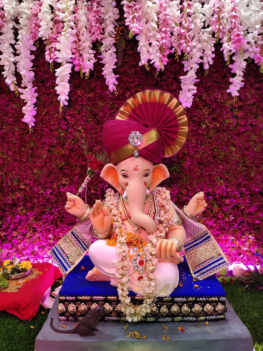 Ganesha deity statue adorned with floral garlands and a traditional turban against a vibrant pink flower wall HD phone wallpaper