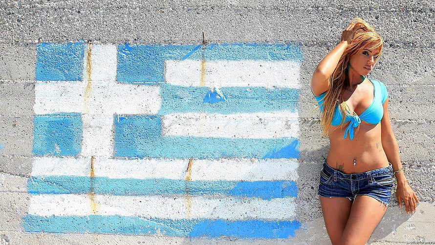 Blonde woman in blue bikini top and denim shorts posing against a painted Greek flag on a concrete wall HD wallpaper