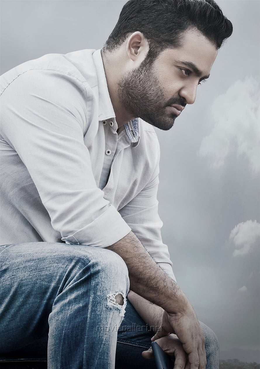 Jr NTR in a contemplative pose wearing a white shirt and distressed denim jeans against a cloudy sky HD phone wallpaper
